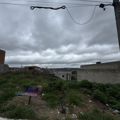 Terra ocupada por mato, poste de energia, céu nublado, prédios ao fundo